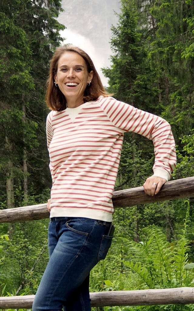 Auf diesem Bild ist eine Frau zusehen die im Wald an einem Holzzaun lehnt und dabei lächelt.